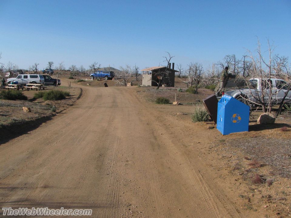 Dirt road leads to a campsite with multiple trucks, a small building, and a blue recycling bin