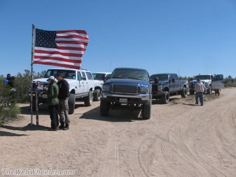 1996 Ford C-350 (Centurion Conversions) - A convoy of trucks and an SUV gathered on a dusty road, with an American flag waving in the breeze.