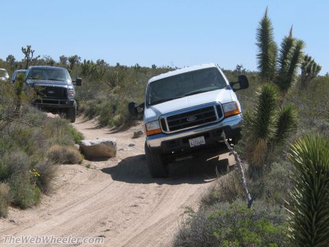 1996 Ford C-350 (Centurion Conversions) - Two Ford F-Series trucks navigate a desert trail with sparse vegetation and clear blue skies.