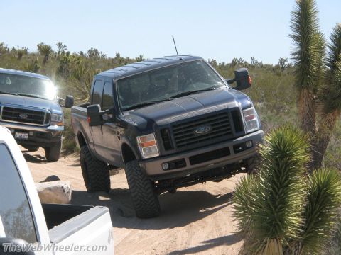 1996 Ford C-350 (Centurion Conversions) - A dark blue Ford F-Series truck navigates a desert trail, with another truck visible in the background.