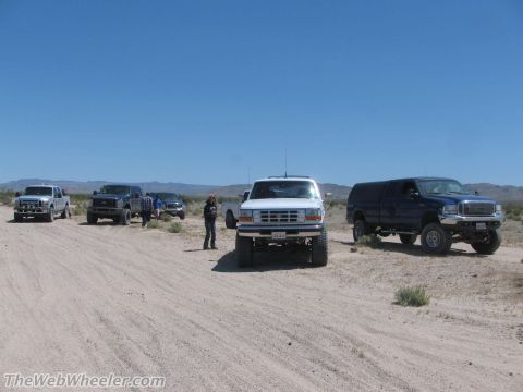 1996 Ford C-350 (Centurion Conversions) - Several lifted trucks, including a 1996 Ford C-350, are parked on a dirt road in a desert area.