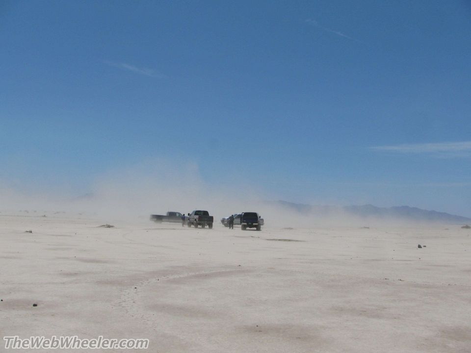 Several trucks driving in a dusty, flat, open area under a clear blue sky