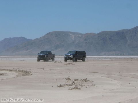 1996 Ford C-350 (Centurion Conversions) - Two trucks traverse a dry lake bed under a clear blue sky, with distant mountains providing a backdrop.
