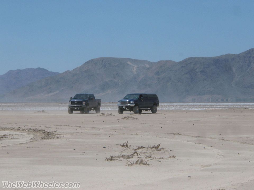 Two dark blue trucks driving on a dry lake bed with mountains in the background. The truck on the left is a pickup, and the truck on the right is an SUV