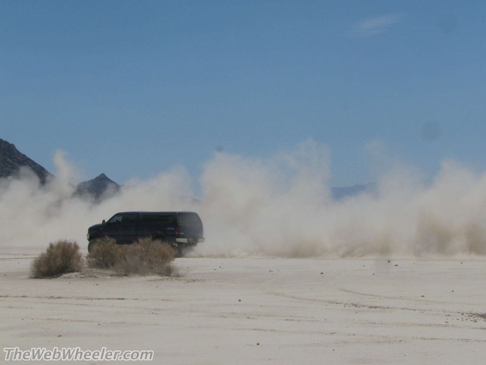 Black SUV driving on a dirt road, kicking up a large dust cloud. Mountains in the background