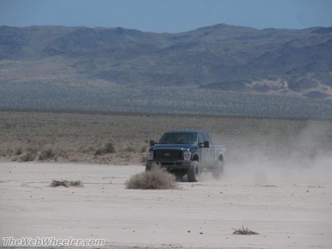 1996 Ford C-350 (Centurion Conversions) - A blue Ford F-Series truck drives through a dusty, arid landscape with mountains in the distance.