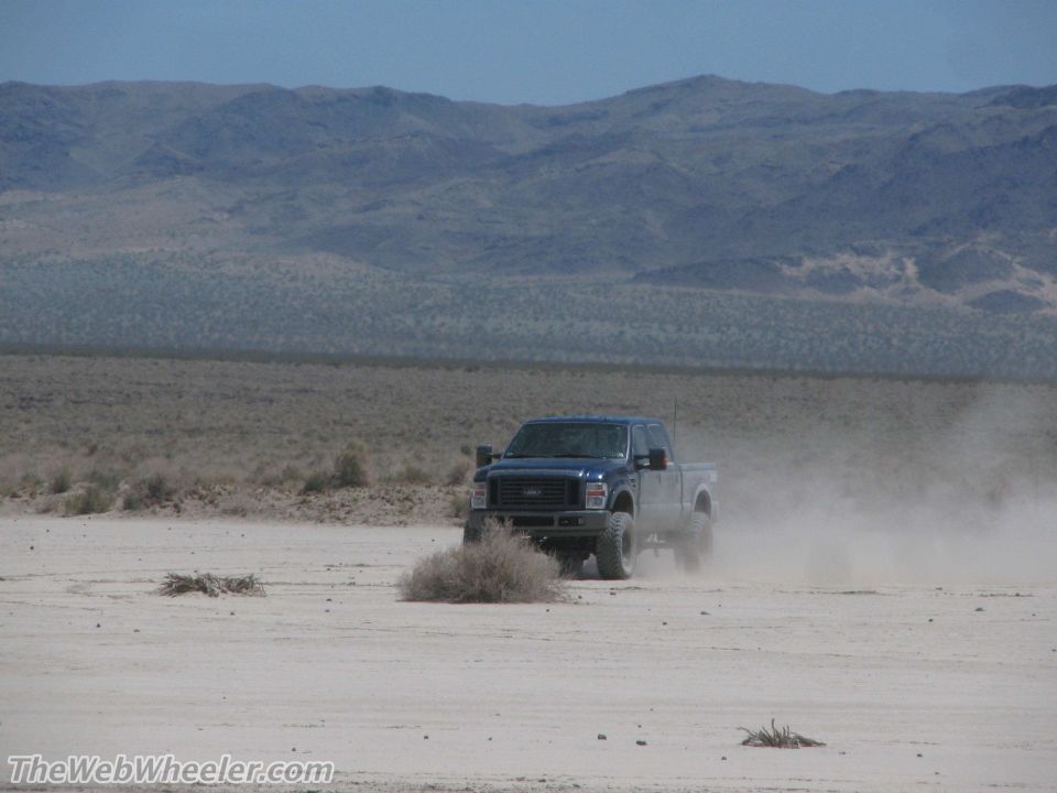 Blue Ford F-Series truck driving on a dirt road, kicking up dust. Mountains in the background