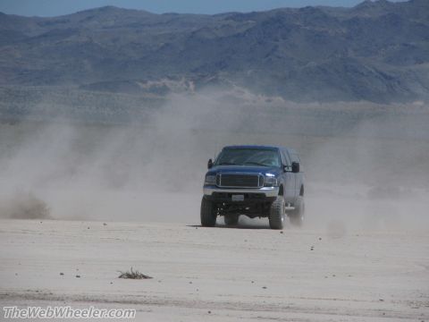 1996 Ford C-350 (Centurion Conversions) - A blue SUV drives through a dusty desert landscape, its tires kicking up a cloud of dirt.