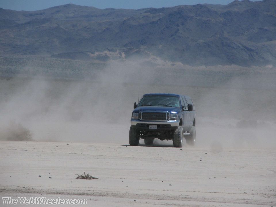 Blue SUV with aftermarket grille driving on dirt road, kicking up dust. License plate visible