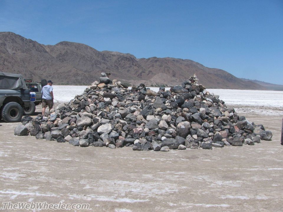 Two rock cairns stand in a dry, flat area with mountains in the background. Two off-road vehicles are parked to the left