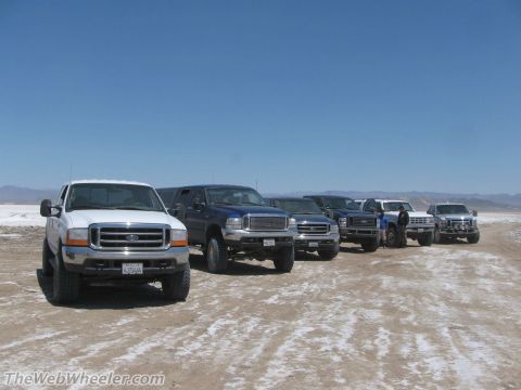 1996 Ford C-350 (Centurion Conversions) - A convoy of Ford Super Duty trucks is lined up on a dusty, white plain with mountains in the distance.