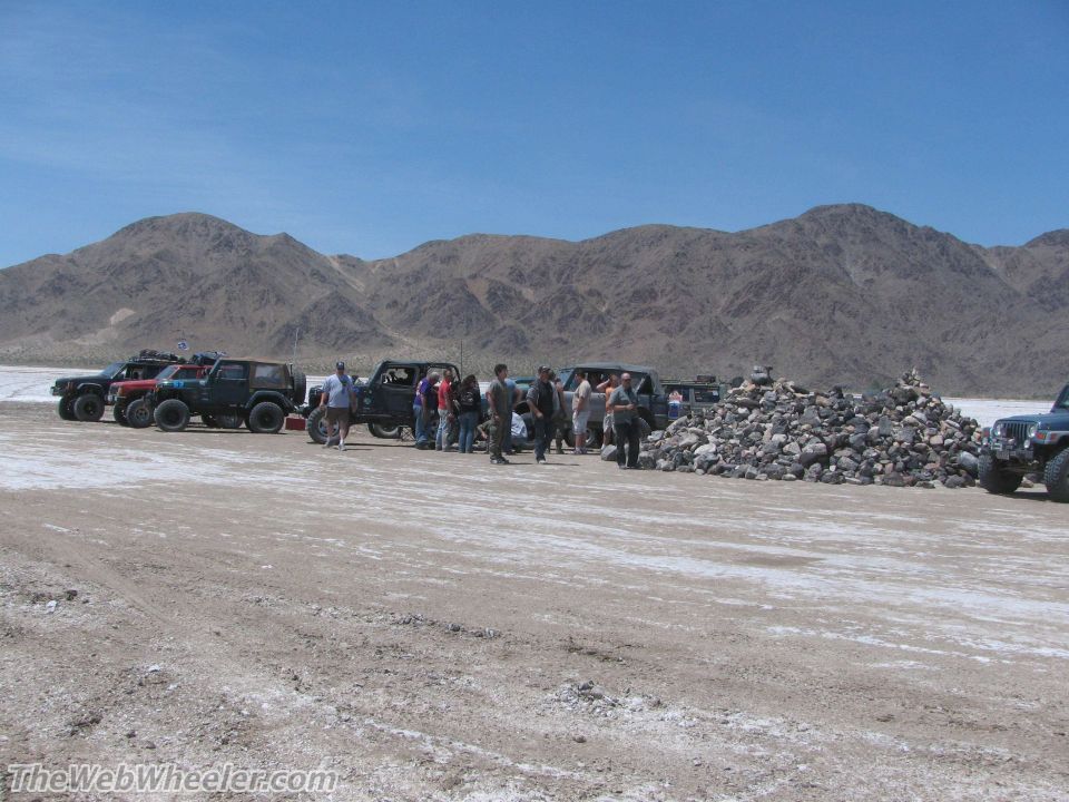Several off-road vehicles parked in a desert with people standing around them and a large pile of rocks nearby