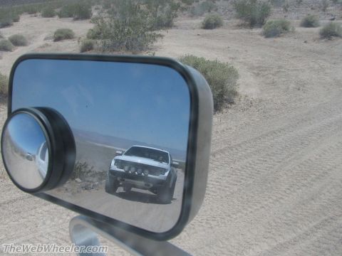 1996 Ford C-350 (Centurion Conversions) - A white pickup truck drives on a dusty desert road, reflected in a side mirror.