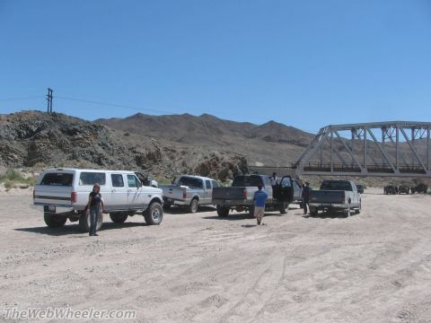 1996 Ford C-350 (Centurion Conversions) - Several pickup trucks and an extended cab SUV are parked near a bridge on a dusty road.