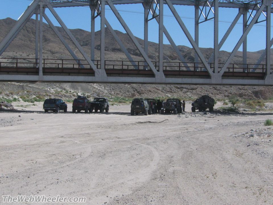 Several off-road vehicles parked on a dirt road under a large metal bridge. Mountains in the background