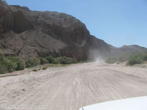 1996 Ford C-350 (Centurion Conversions) - A vehicle kicks up dust on a dirt road through a desert canyon with rocky hills and sparse vegetation.