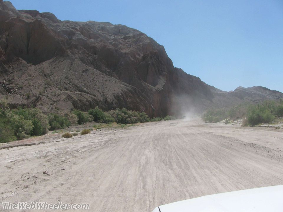 White vehicle hood in foreground, dirt road with dust cloud ahead in a desert canyon