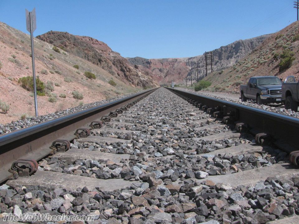 Railroad tracks stretch into the distance through a desert canyon with two trucks parked nearby