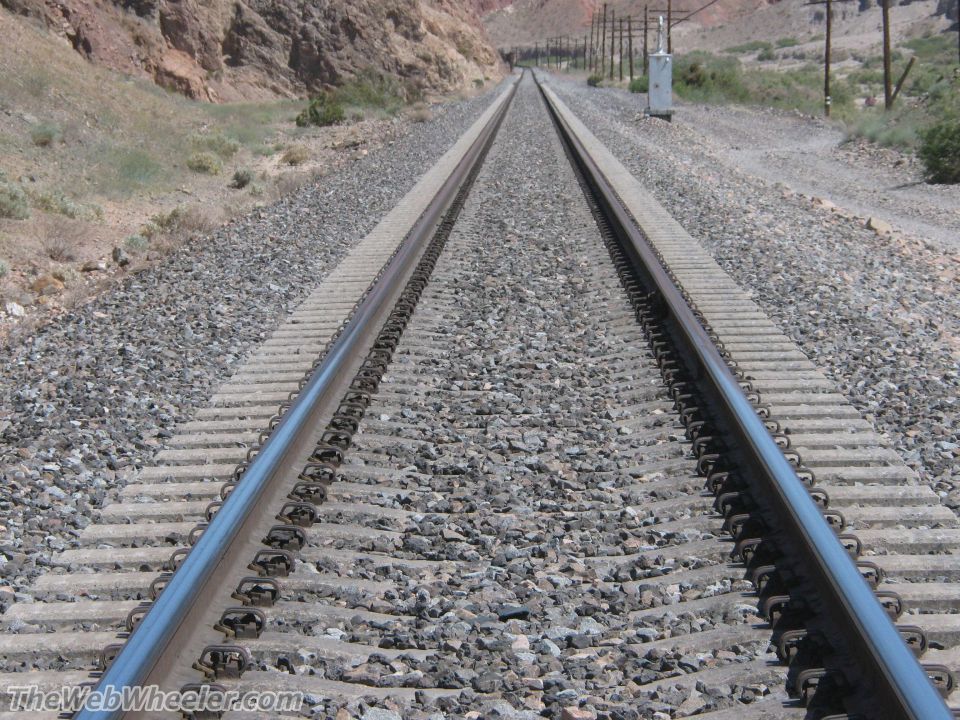 Railroad tracks stretch into the distance through a rocky, arid landscape with sparse vegetation and utility poles