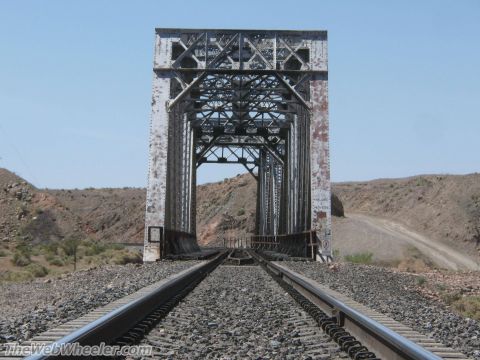 1996 Ford C-350 (Centurion Conversions) - A weathered railroad bridge spans across train tracks in a dry, arid environment under a clear blue sky.