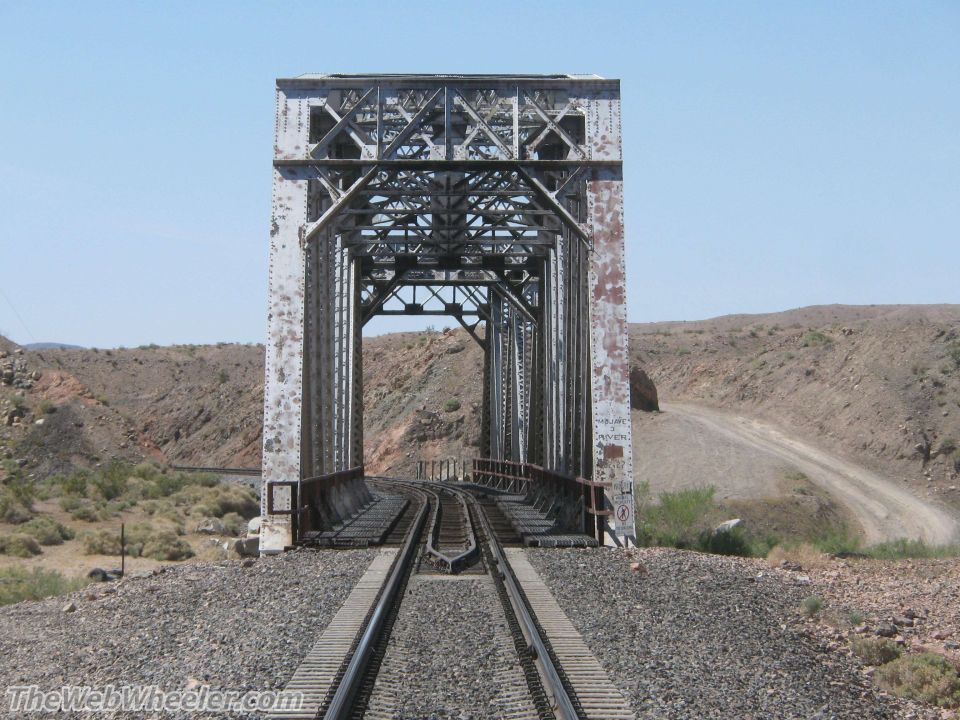 Railroad bridge with steel trusses crossing a desert landscape with a dirt road visible