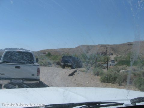 1996 Ford C-350 (Centurion Conversions) - View out through a dirty windshield over a white hood on a desert trail, with a silver Chevrolet pickup parked at left and a dark lifted SU…