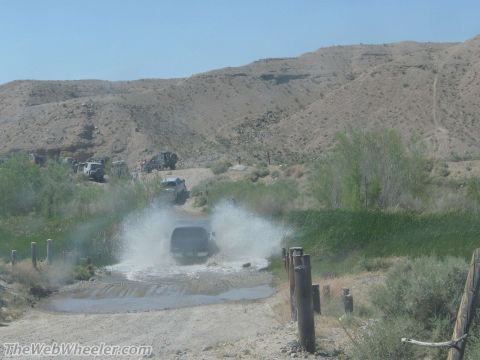 1996 Ford C-350 (Centurion Conversions) - Several off-road vehicles navigate a dusty trail, with one SUV kicking up a large spray as it crosses a shallow stream.