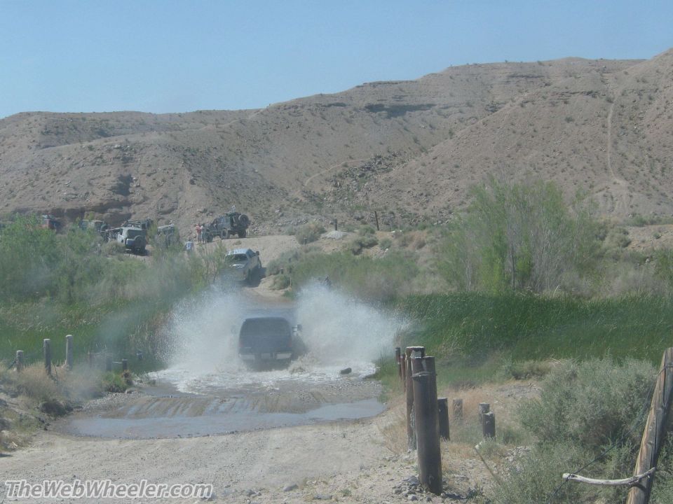 Dark SUV splashing through water crossing, other vehicles in background