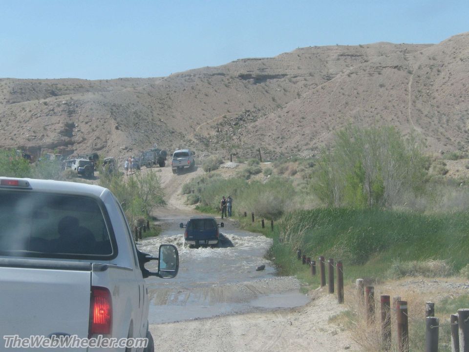 Several vehicles driving through a shallow water crossing on a dirt road with hills in the background