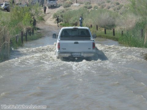 1996 Ford C-350 (Centurion Conversions) - A white pickup truck navigates through a flooded dirt road, with several people and other vehicles visible on the higher ground.