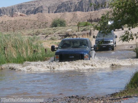1996 Ford C-350 (Centurion Conversions) - Two Ford F-Series trucks navigate a shallow water crossing in a dry, mountainous landscape.