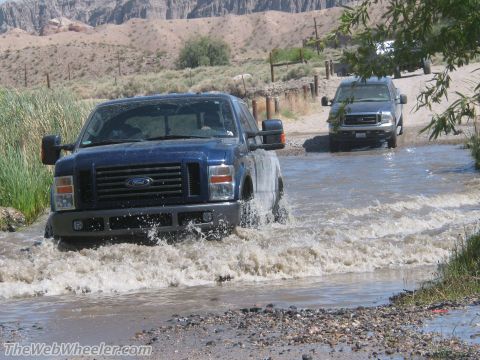 1996 Ford C-350 (Centurion Conversions) - Two Ford F-Series trucks navigate a shallow water crossing on a dusty trail.