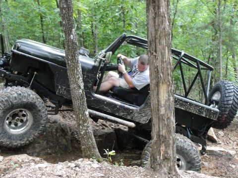 1967 Ford Mustang - A black off-road jeep with a roll cage is driven through a wooded area with large rocks.
