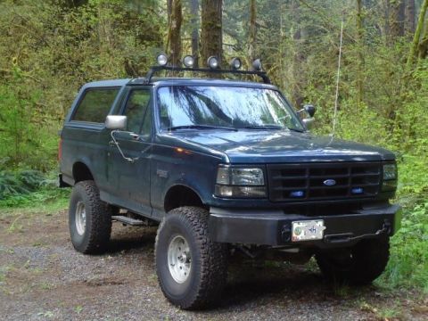 1995 Ford Bronco - This lifted Ford Bronco sits on a gravel path surrounded by trees, equipped with auxiliary lights on its roof rack.