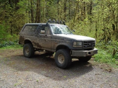 1995 Ford Bronco - This 1995 Ford Bronco, seen in a wooded area, is heavily covered in mud and features auxiliary lights on its roof rack.