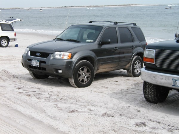 Dark gray Ford Escape parked on beach sand with roof rails, water and boats behind