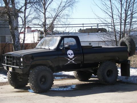 1990 Chevrolet Suburban - This lifted black pickup truck features a prominent skull and crossbones decal on the door and a spare tire mounted at the rear.