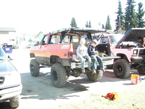 1990 Chevrolet Suburban - Two boys sit on the tailgate of a lifted orange SUV next to a pink truck with its hood open.