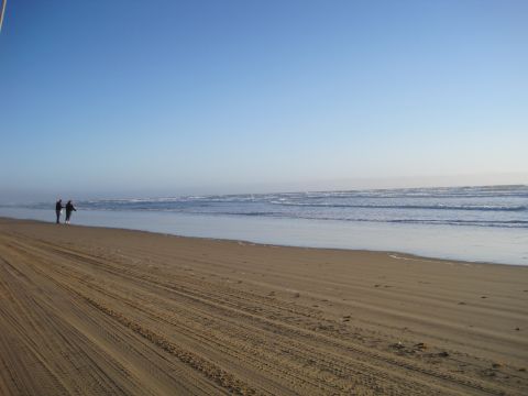 1989 Toyota Pickup/SR5 - A wide shot of a beach with tire tracks in the sand and two figures walking near the water.