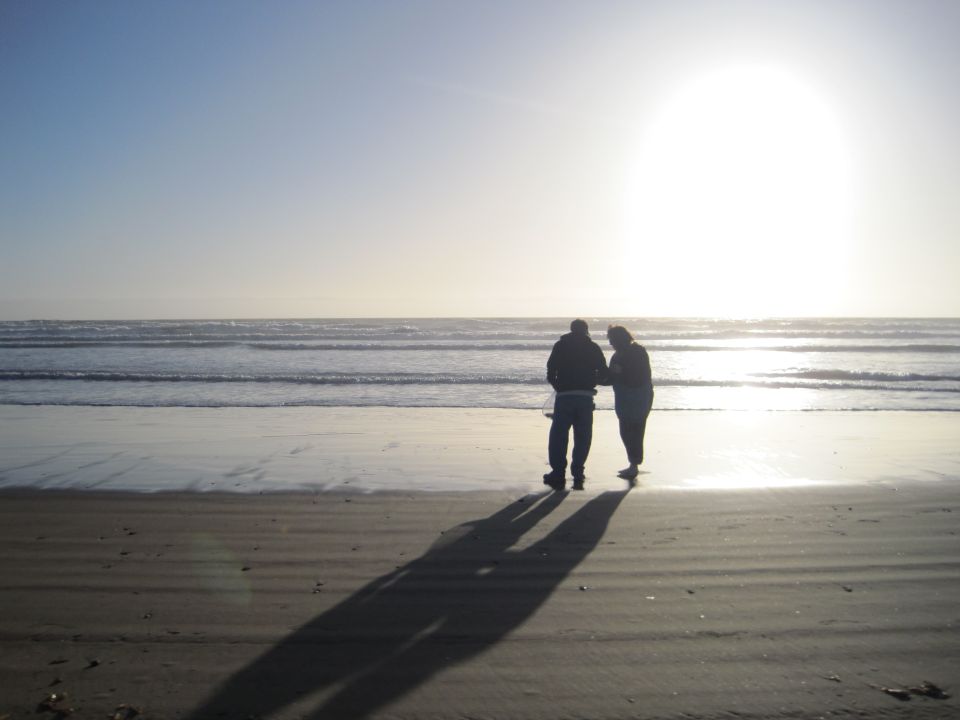 Two people walk on a sandy beach with ocean waves and a bright sun