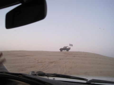 1989 Toyota Pickup/SR5 - A dark SUV flies a checkered flag while cresting a sand dune under a hazy sky.