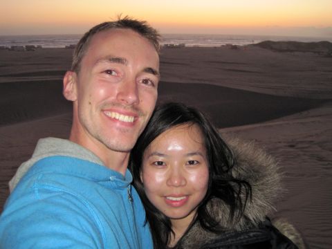1989 Toyota Pickup/SR5 - A couple poses for a selfie on sand dunes with the ocean visible in the distance during sunset.