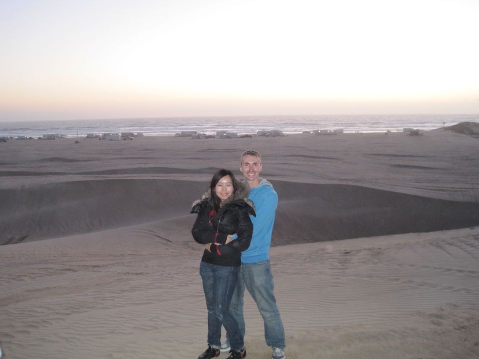 Couple posing on sand dunes with the ocean and distant RVs in the background