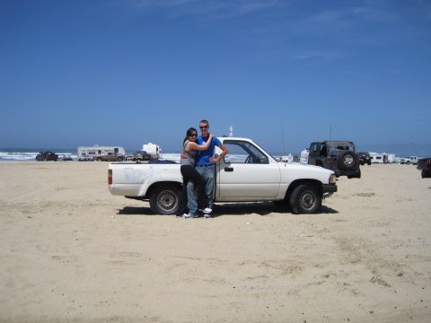 1989 Toyota Pickup/SR5 - A couple stands in front of a white 1989 Toyota Pickup/SR5 on a sunny beach day with other vehicles and RVs in the background.
