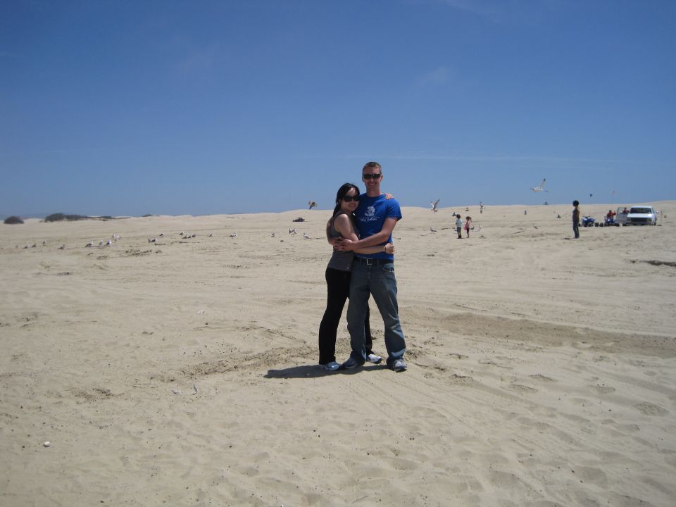 Couple hugging on a beach with birds and a white SUV in the distance