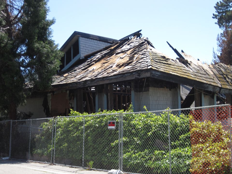Damaged house with a partially collapsed roof behind a chain-link fence
