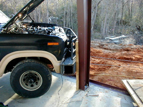 Black square-body truck with hood up parked at a concrete pad beside a steel I-beam and fresh red dirt excavation