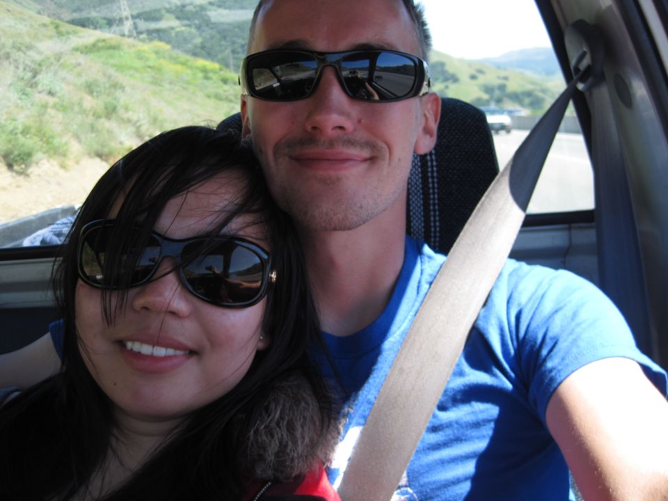 Two people in sunglasses taking a selfie inside a vehicle with hills outside the window