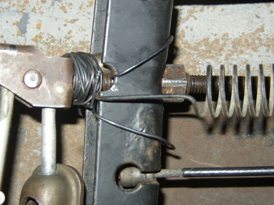 Close-up of linkage with threaded rod, return spring, wire-wrapped lever and cable on a black bracket, rusty background