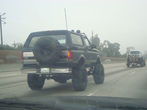 1986 Ford Bronco - A heavily modified Ford Bronco with oversized tires drives down a hazy highway, with another vehicle in the distance.